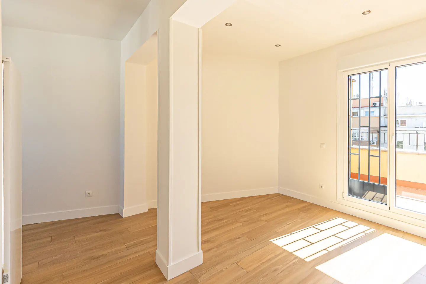 Bright, empty room with light wood floors, white walls, and a large window with bars. Sunlight streams in, creating shadows on the floor.