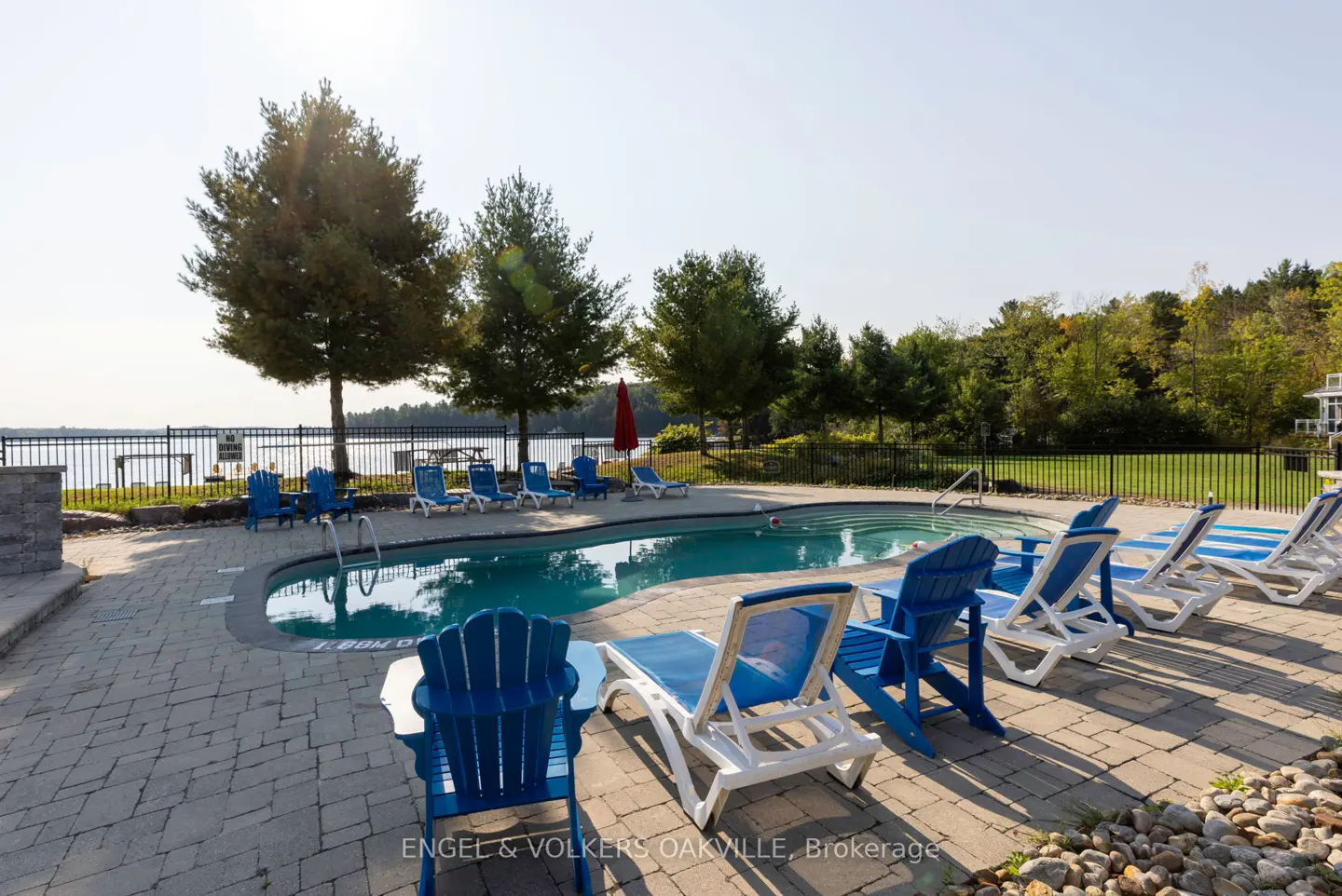 Outdoor pool with blue and white lounge chairs on a brick patio. Trees and a lake are visible in the background.