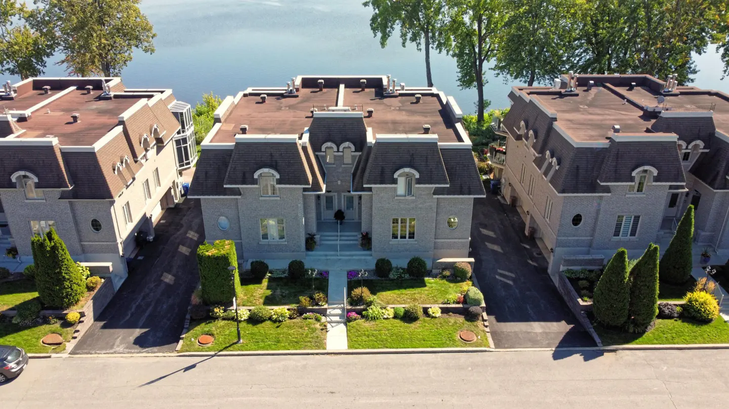 Aerial view of three grey stone townhouses with brown roofs, green lawns, and a lake in the background.