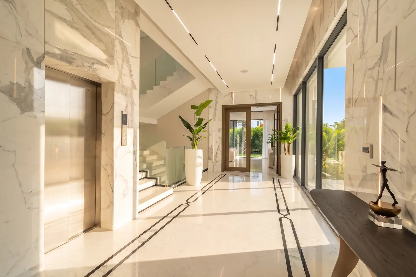 Bright, modern foyer with marble walls and floors. An elevator is on the left, stairs in the back, and a table with a sculpture on the right.