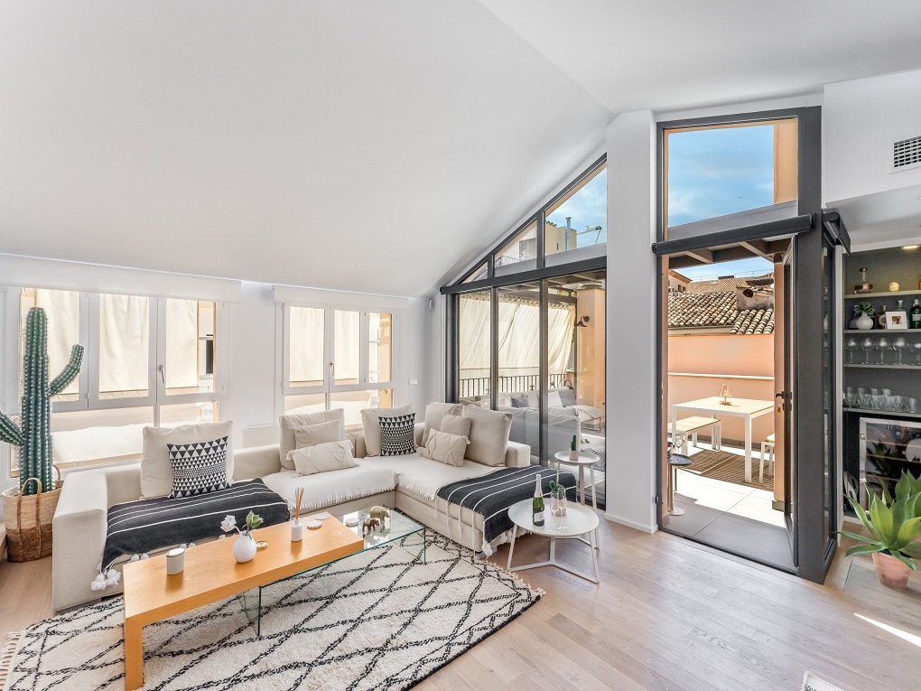 Bright living room with a large white sectional, wooden coffee table, patterned rug, and glass doors opening to a terrace. [AI-generated text]