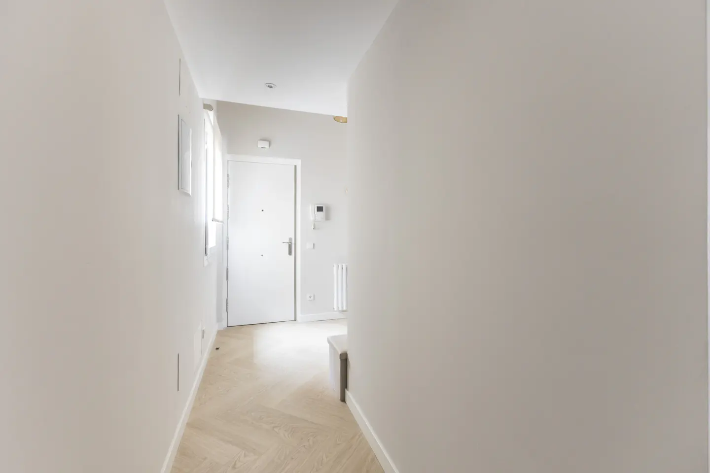 Hallway with light wood floors, white walls, and a white door at the end. A small bench is visible on the right.