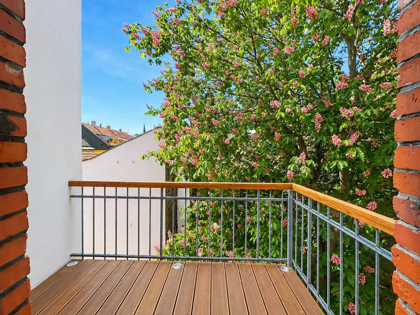 Balcony view with wood floor, metal railing, and brick walls. A tree with pink flowers is visible in the background against a blue sky.