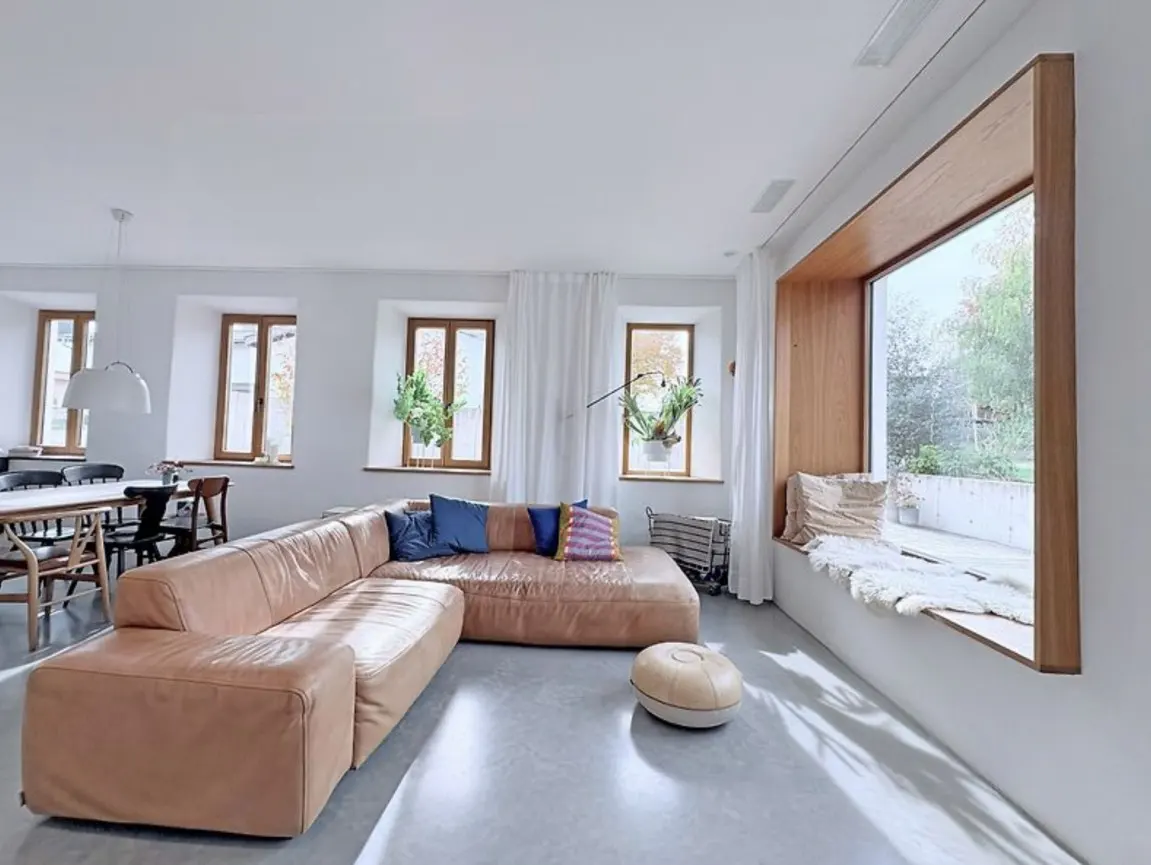 Bright living room with a tan leather sectional sofa, blue pillows, and a window seat. White walls and concrete floors create a modern, airy space.