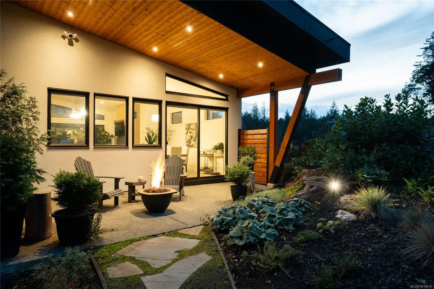 Outdoor patio with a fire pit, chairs, and lush landscaping. The modern home has a wood ceiling and black framed windows.