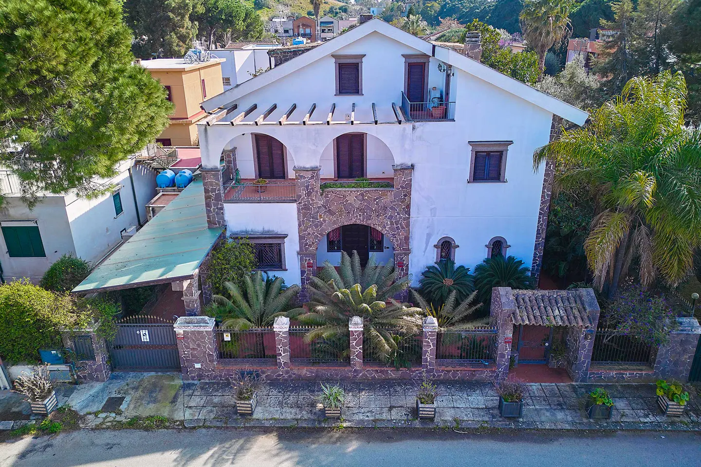 Two-story white house with stone accents, dark brown shutters, and a stone fence with tropical plants.