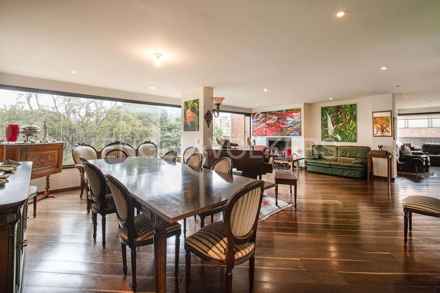 Spacious dining room with a long wooden table, chairs, and hardwood floors. Large windows offer a view of lush greenery. Art adorns the walls.