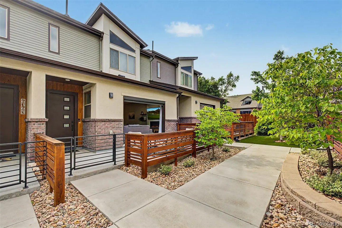 Exterior of a modern townhouse with a brown door, brick accents, and a wooden fence. A concrete walkway leads to the entrance.