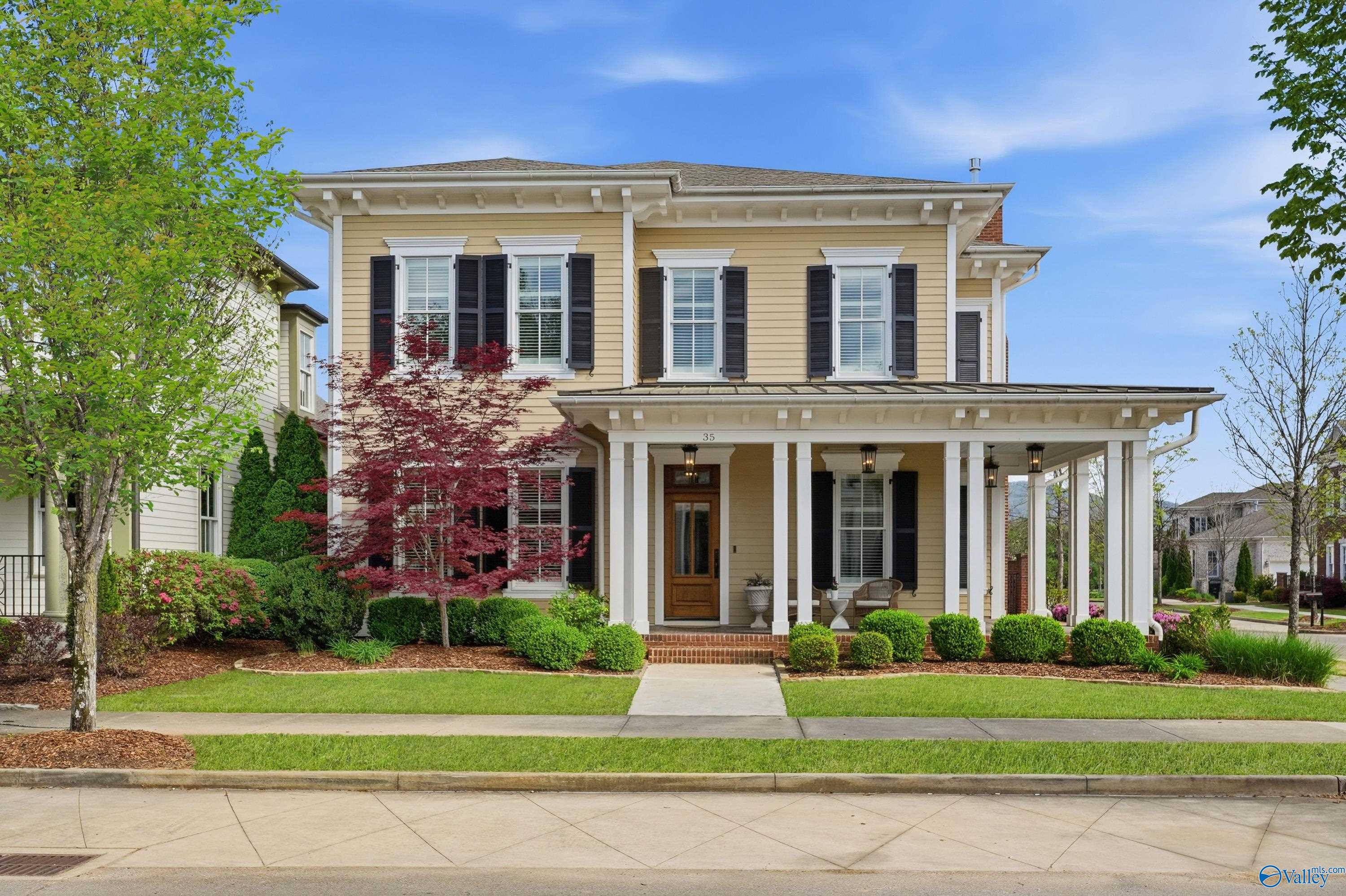 Two-story yellow house with black shutters, white trim, and a covered front porch with white columns. A green lawn and trees surround the house.