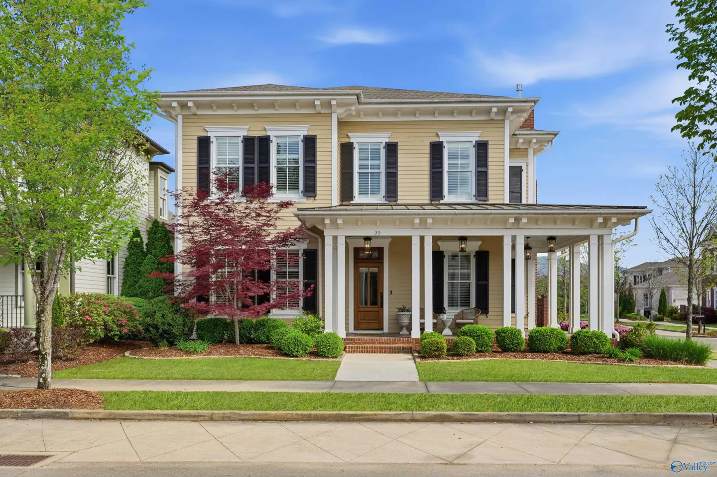 Two-story yellow house with black shutters, white trim, and a covered front porch with white columns. A green lawn and trees surround the house.