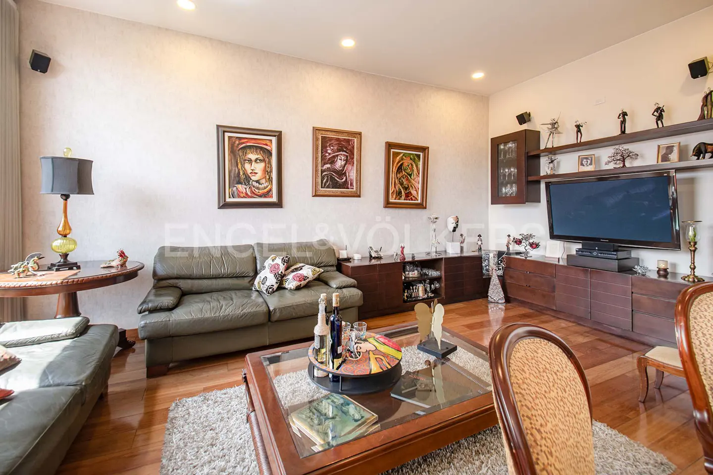 Living room with hardwood floors, a green leather sofa, a glass-topped coffee table, and a large television. Three framed portraits hang on the wall.