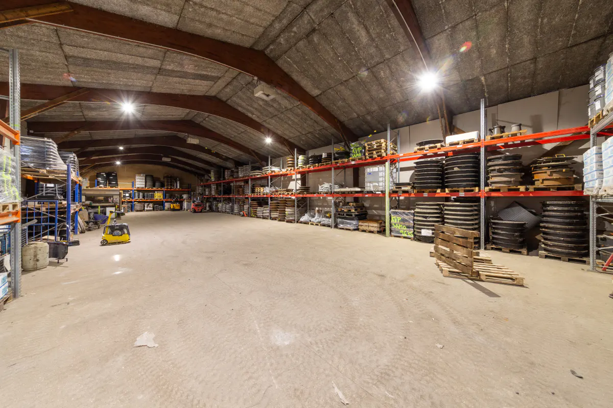 A wide shot of a large warehouse with high ceilings, metal shelving, and a concrete floor. Pallets and various items are stored on the shelves.