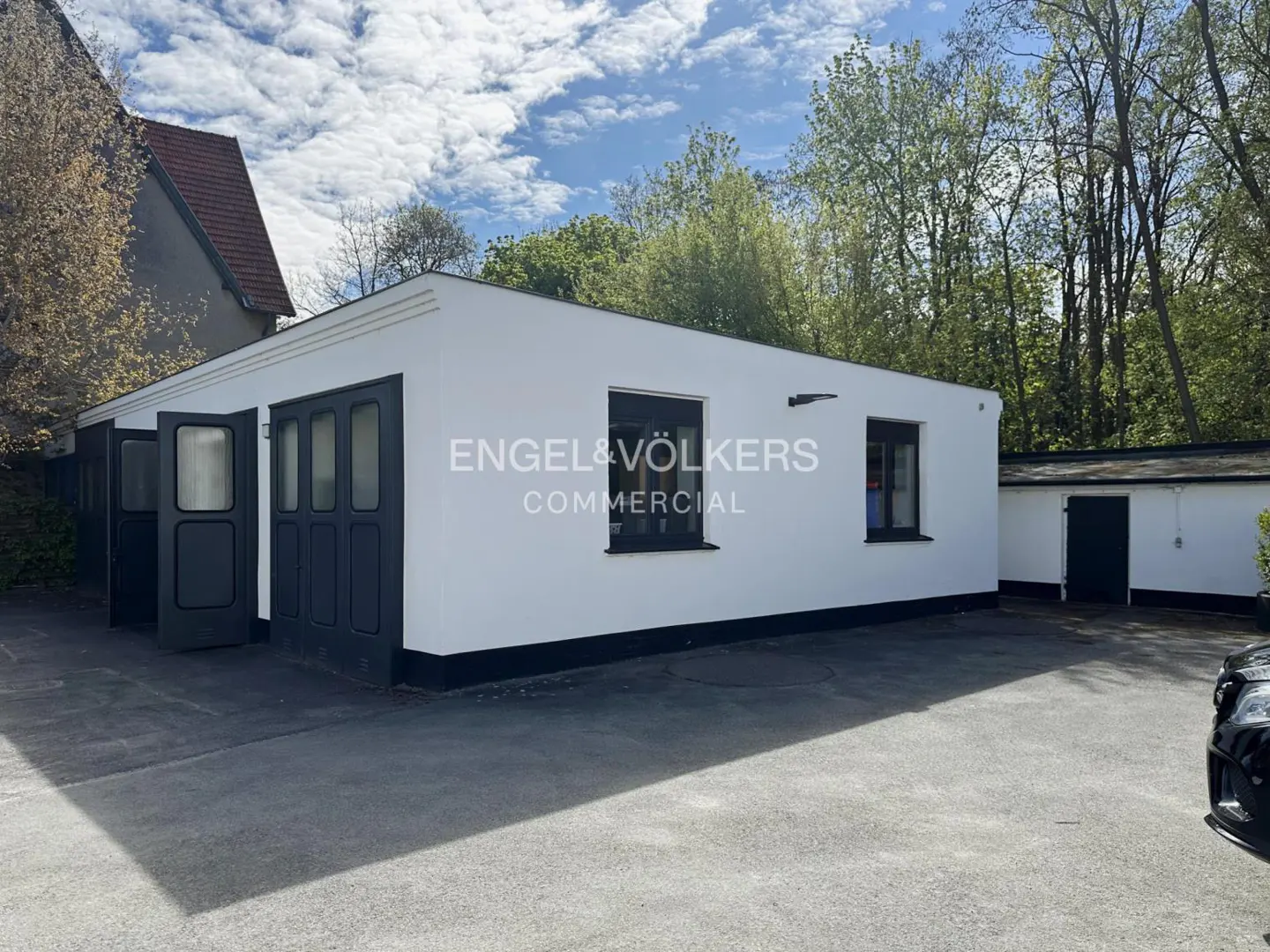 Exterior of a white commercial building with black trim and garage doors, Engel & Völkers logo, and a paved parking area.