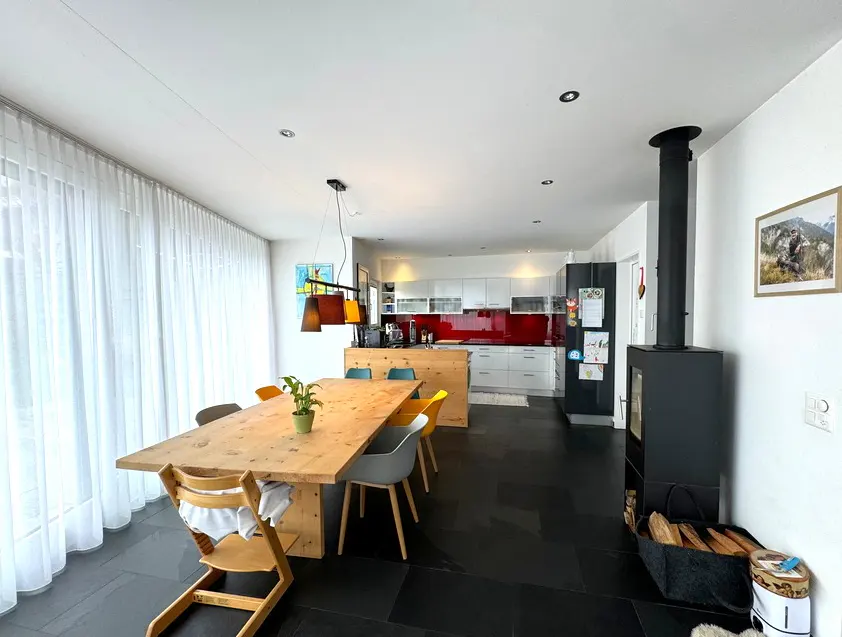 Bright, open-concept living space with a wood dining table, modern chairs, and a black wood-burning stove. White kitchen cabinets with red backsplash in background.