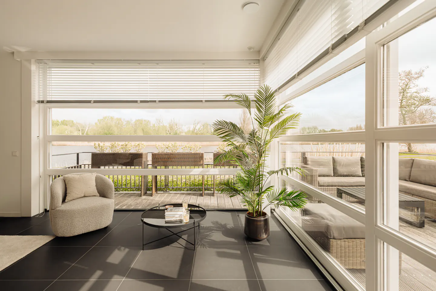 Bright sunroom with black tile floor, large windows, and white blinds. A potted palm sits near the open door to a deck with outdoor seating.