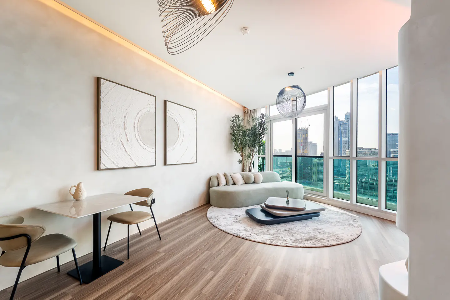 Bright, modern living room with wood floors, a light green sofa, and a round rug. Large windows offer a city view. Two abstract art pieces hang on the wall.