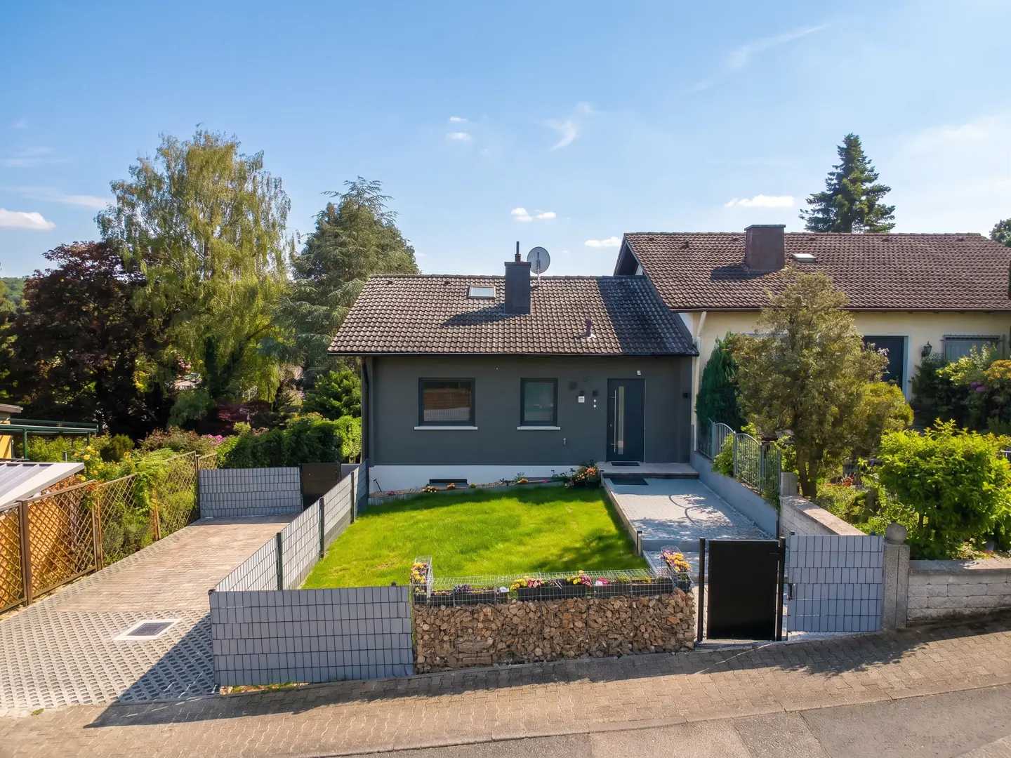 A grey house with a green lawn, a stone-filled planter, and a paved driveway behind a modern fence under a blue sky.