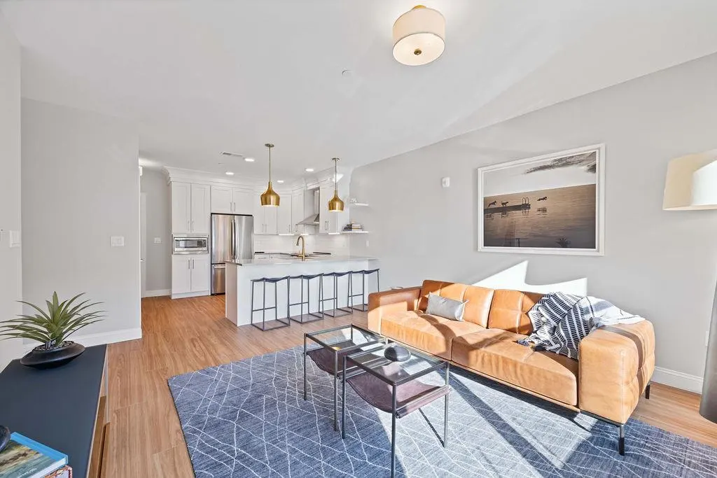 Bright, open-concept living space with a tan leather sofa, blue rug, and white kitchen with island seating.