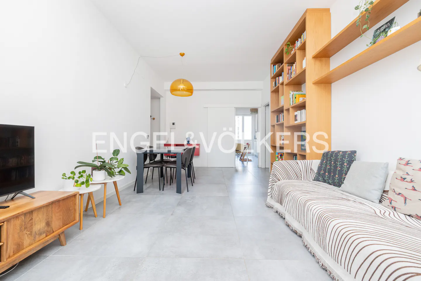 Bright living room with gray tile floor, dining table, sofa, and wooden bookcase. A yellow pendant light hangs above the table.