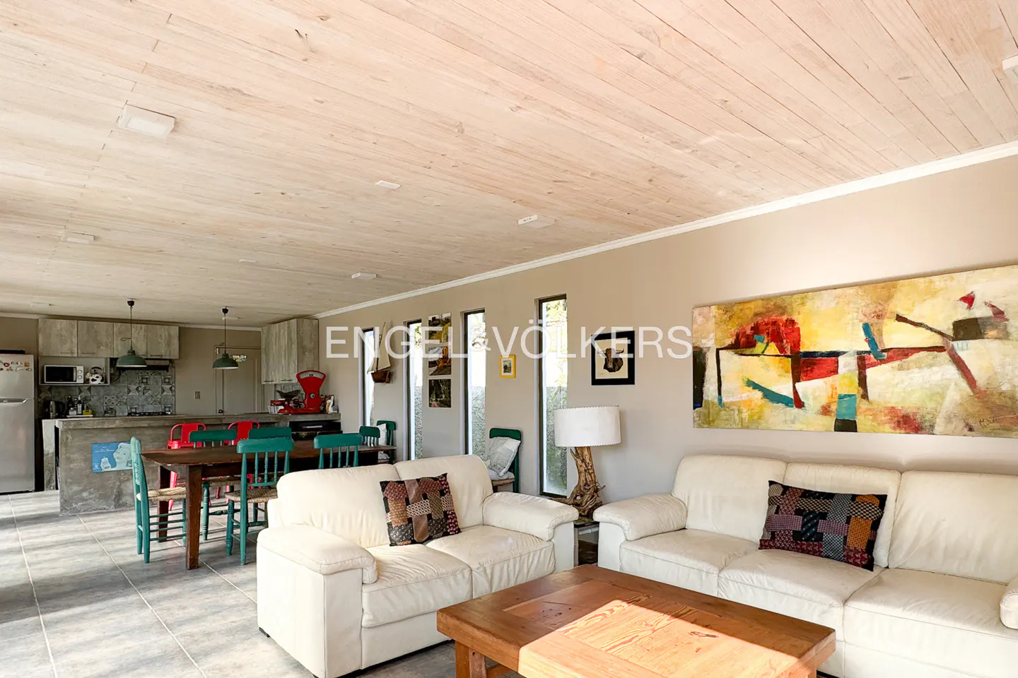 Open-plan living area with white sofas, wooden coffee table, dining set, and large abstract painting on a beige wall.