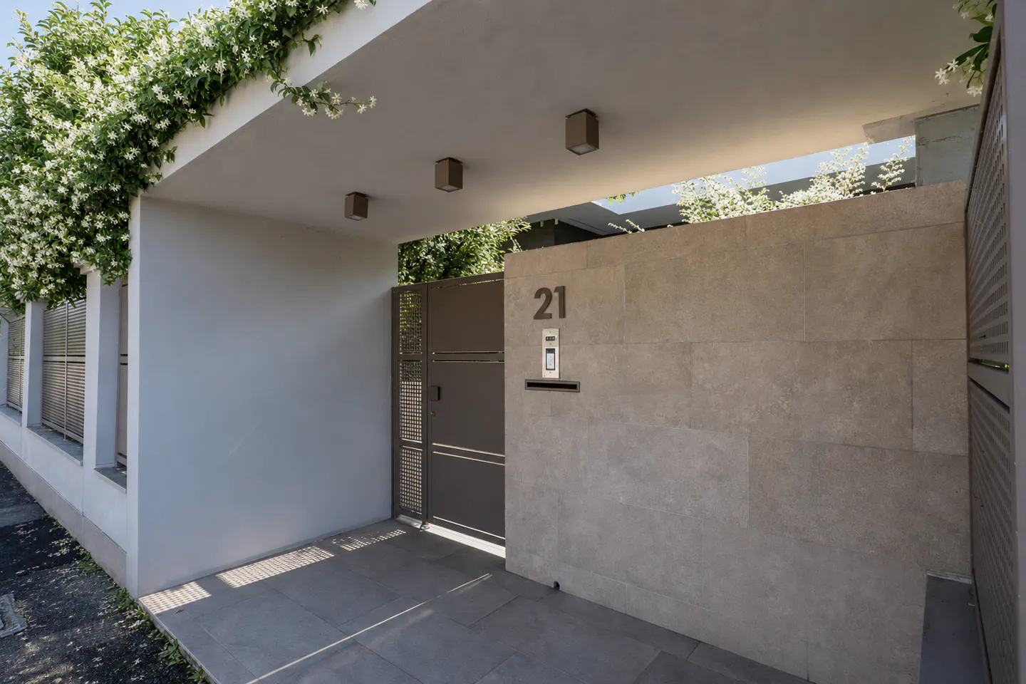 Modern home entrance with gray walls, a metal gate, and the number 21 above a doorbell and mail slot. White flowers adorn the top.