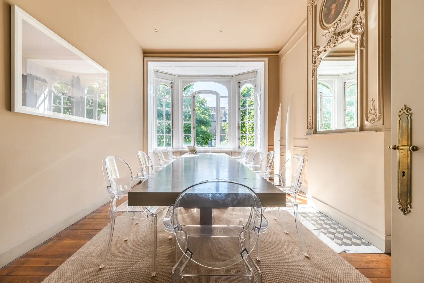 A dining room with a long table, clear chairs, and a bay window. A large framed picture hangs on the wall. A mirror is on the right wall.
