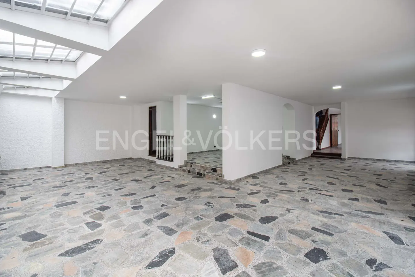 Spacious room with stone floors, white walls, and a skylight. Engel & Völkers logo visible. Stairs lead to another room.
