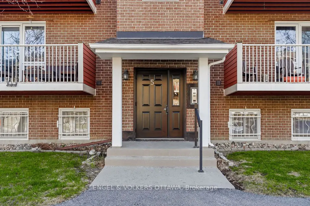 Brick apartment building with brown door under a white portico, number 5 above the door. Balconies with white railings on the second floor.
