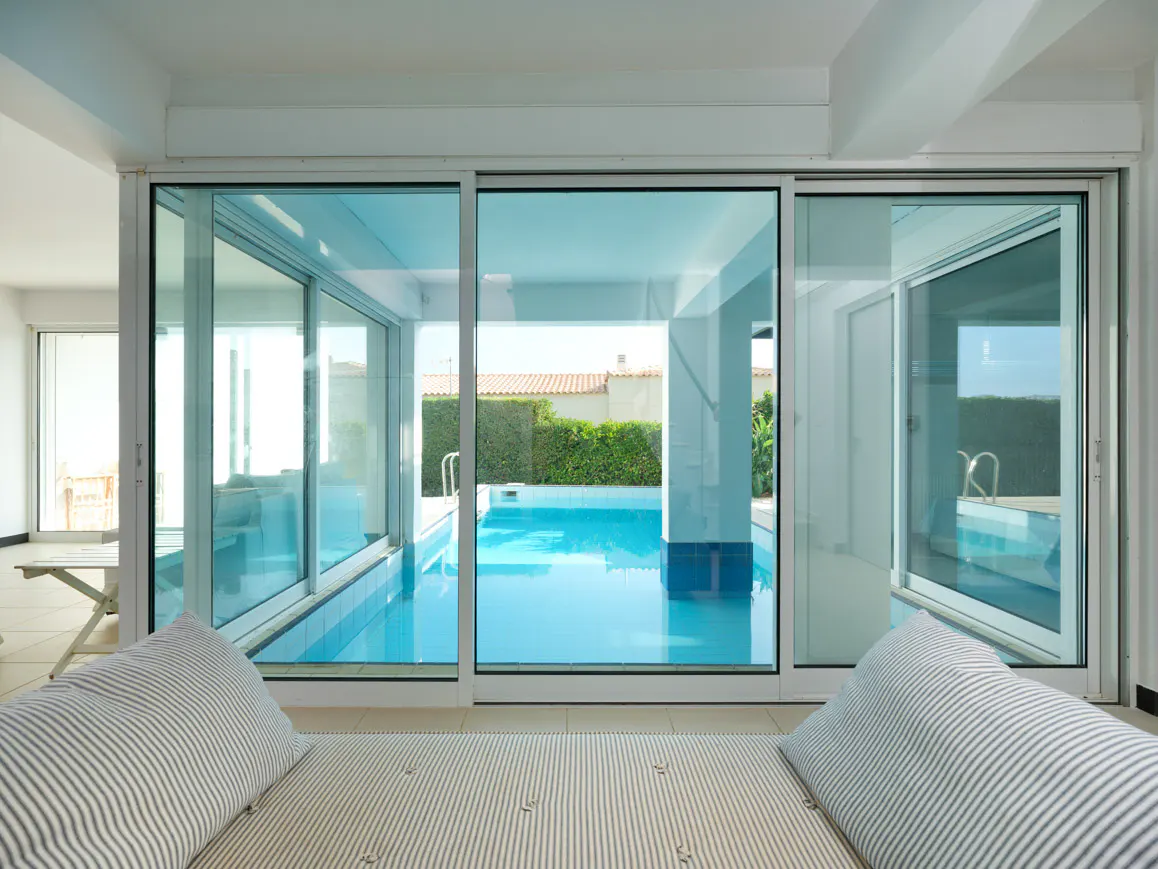 View from a room with striped cushions through large glass sliding doors to a bright blue outdoor swimming pool.