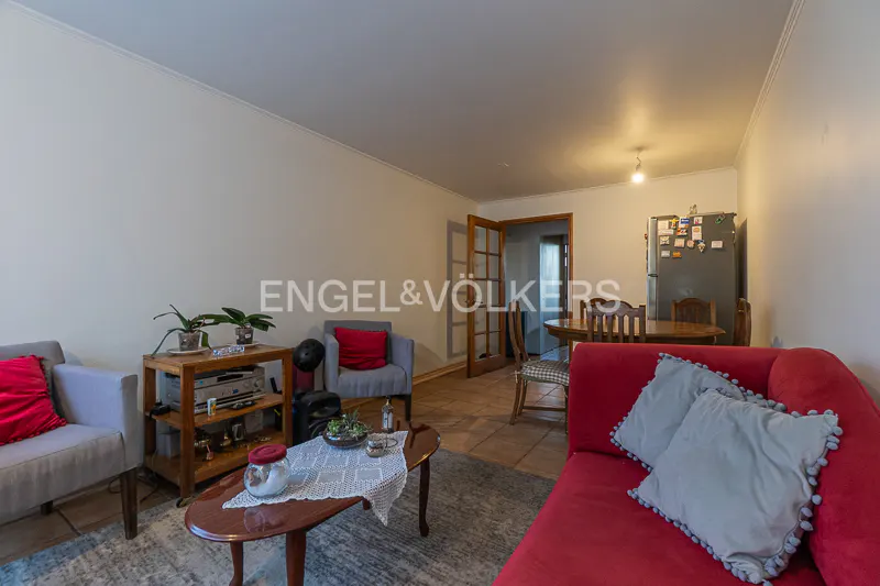 Living room with red sofa, gray chairs, and wooden table. Kitchen with fridge and dining table in the background.