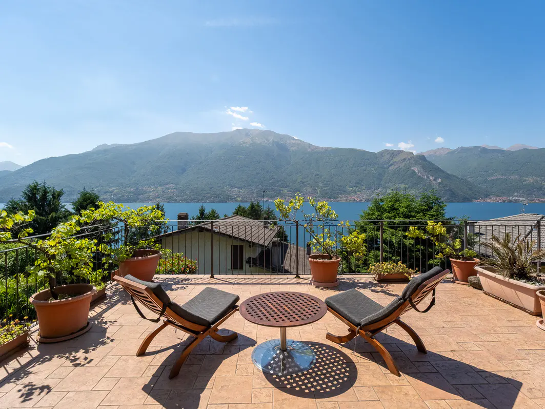 A sunny terrace with two wooden chairs and a round table, overlooking a lake and mountains under a clear blue sky.