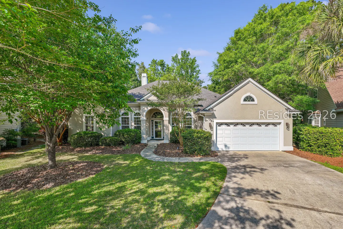 Beige single-story house with a white garage door, arched entryway, green lawn, and trees under a blue sky.