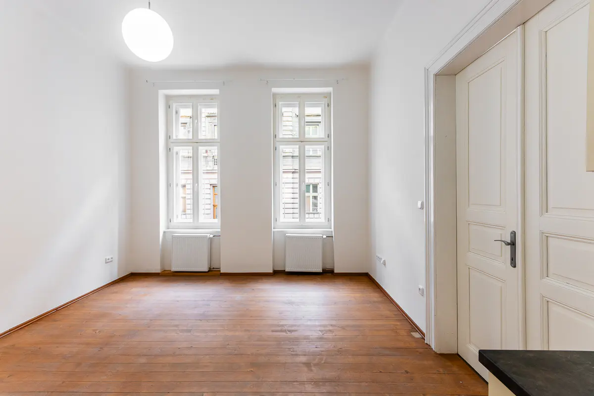 Bright, empty room with hardwood floors, white walls, and two windows. A white door is on the right.