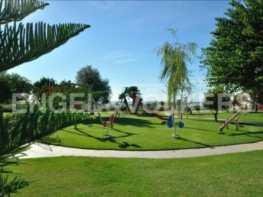 A playground with green grass, trees, and blue sky. There is a slide, spring riders, and a seesaw.