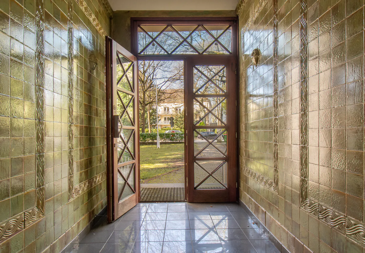 Entryway with glossy olive-green tiled walls and a wooden door opening to a sunny garden view.