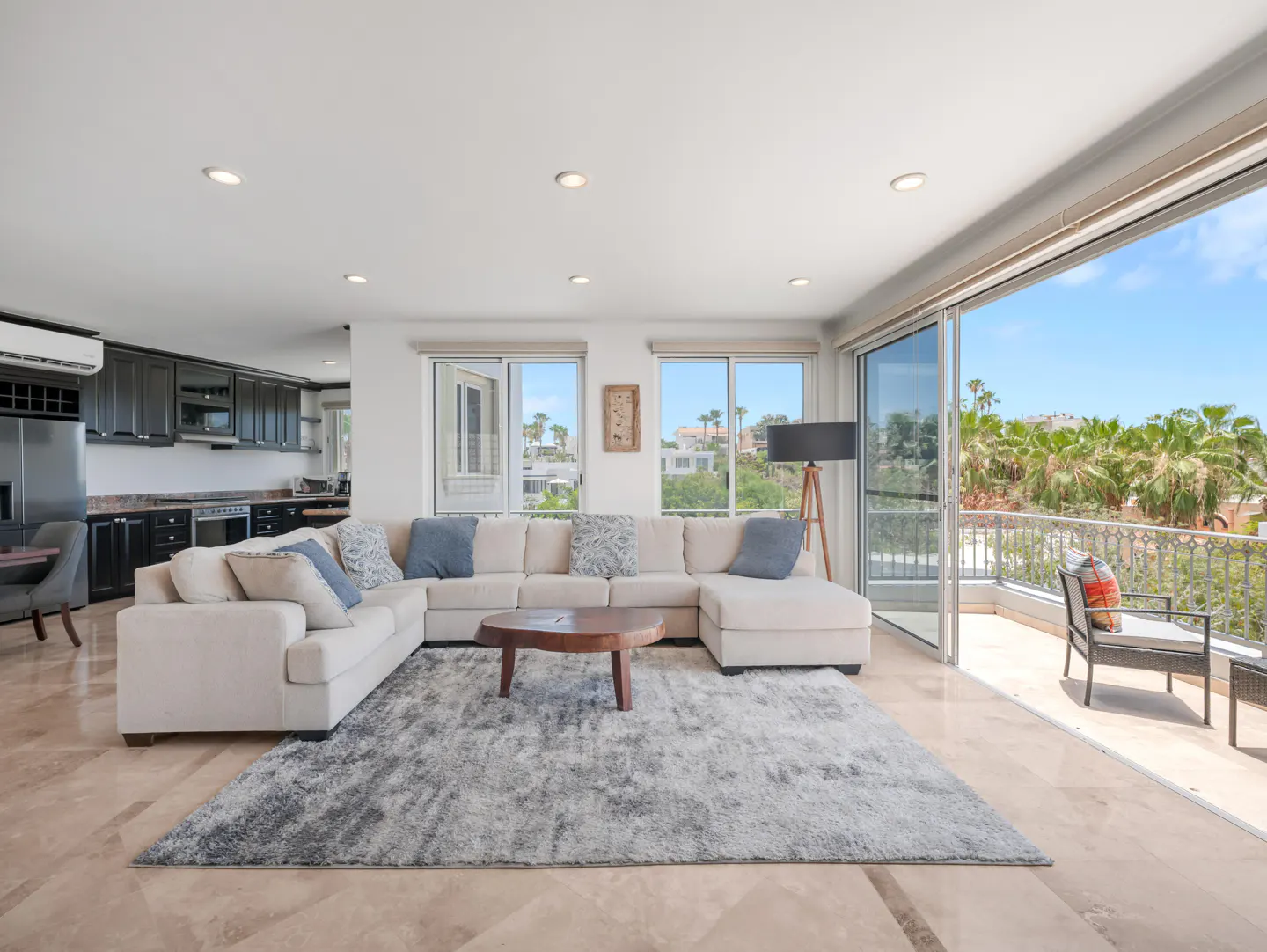 Modern living room with a large beige sectional, grey rug, and open sliding doors leading to a balcony with palm trees.