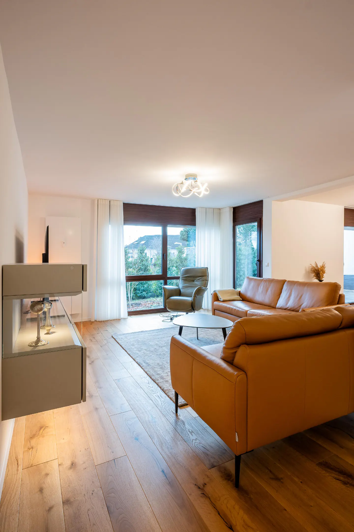 Bright living room with hardwood floors, tan leather sectional sofa, and large windows with sheer white curtains.