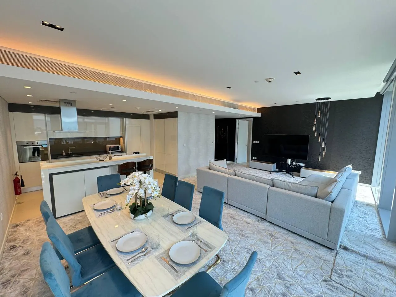 Open-concept living space with a kitchen, dining table with blue chairs, and a gray sectional sofa. A large TV is mounted on a black wall.