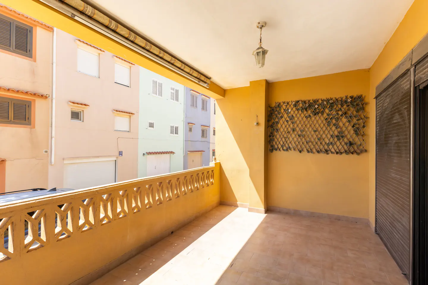 A sunny, yellow balcony with a decorative railing overlooks colorful buildings. A lattice with leaves hangs on the wall.
