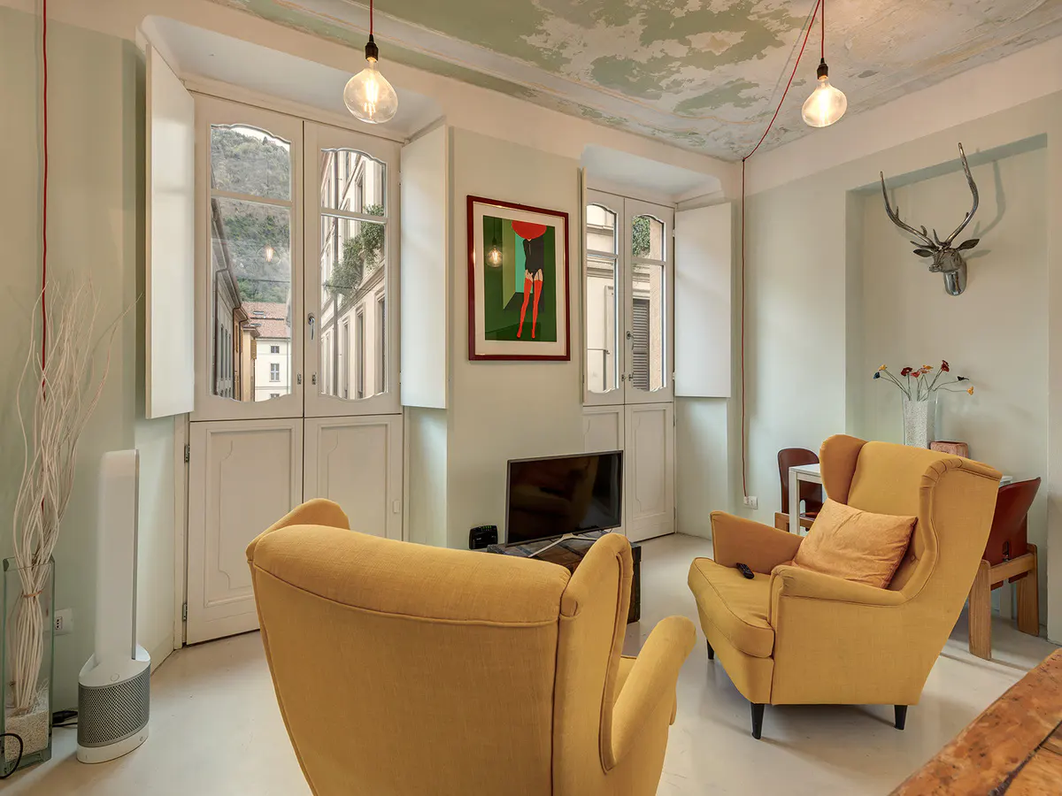 Living room with yellow armchairs, white walls, and tall windows. A deer head mount hangs on the wall. Modern art and pendant lights add style.