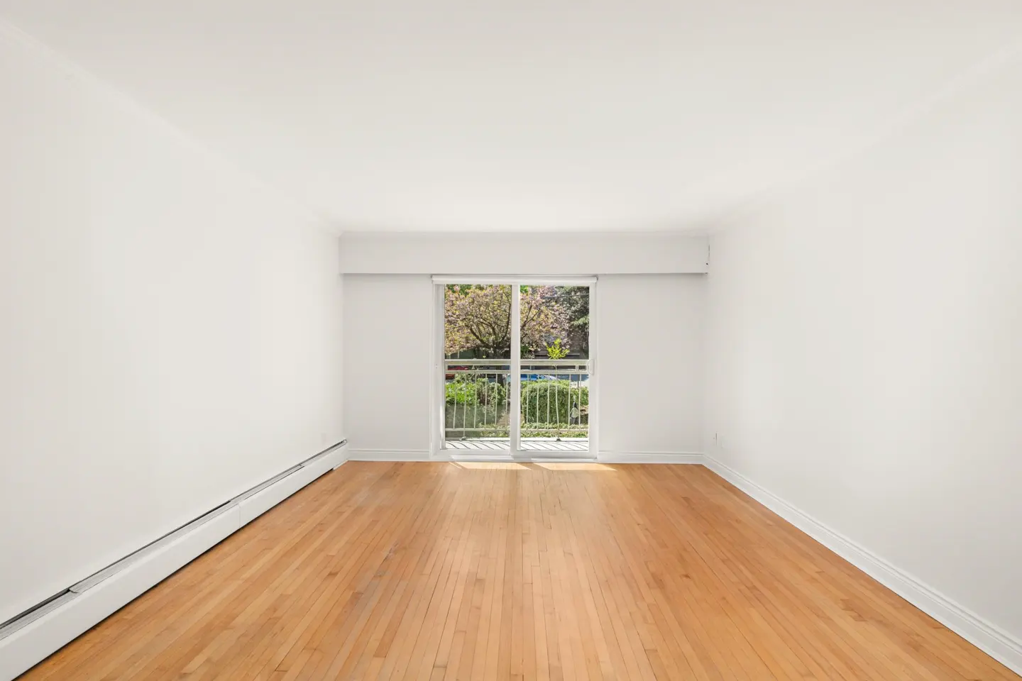 Bright, empty room with hardwood floors and white walls. Sliding glass doors lead to a balcony with a view of trees.