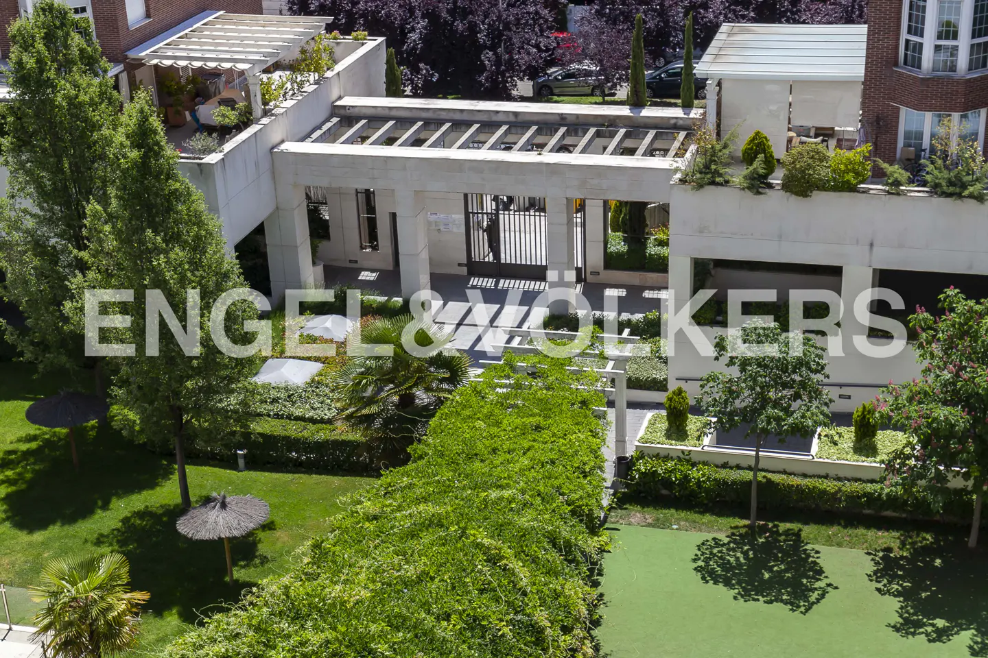 Exterior view of a gated community with lush green landscaping and modern architecture. The Engel & Völkers logo is superimposed on the image.