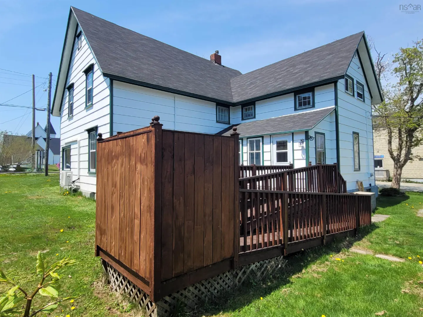 Exterior view of a two-story white house with a dark roof and a brown wooden deck with railings. Green grass surrounds the house.