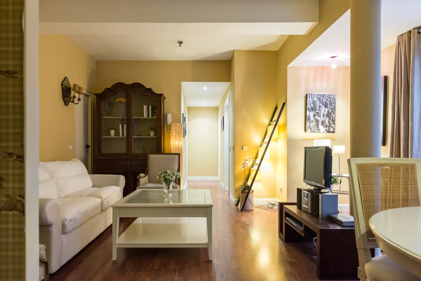 A warm-toned living room features a white sofa, glass-topped coffee table, and a dark wood cabinet. A hallway leads to another room.