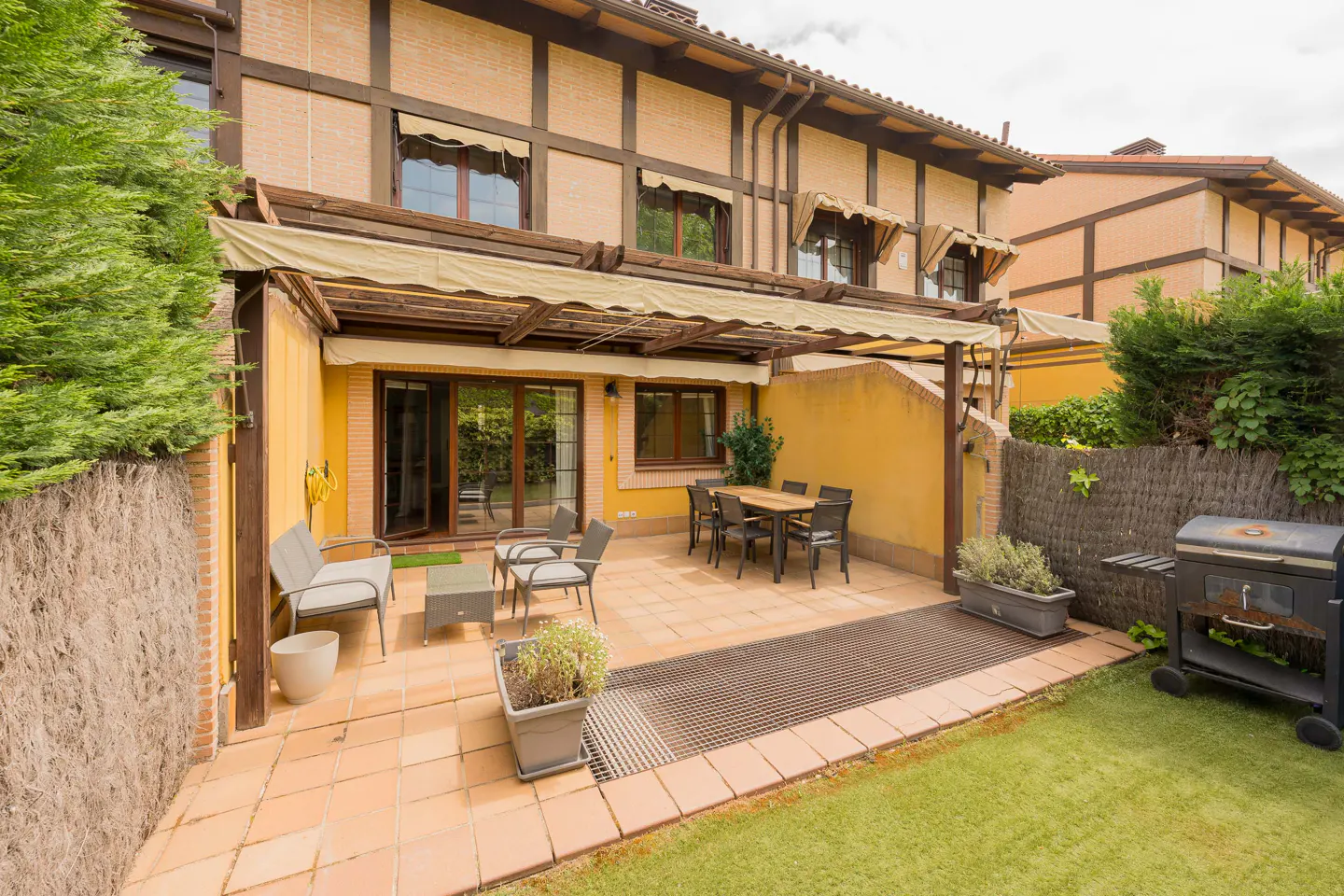 A backyard patio with a table, chairs, grill, and a yellow wall under a pergola.