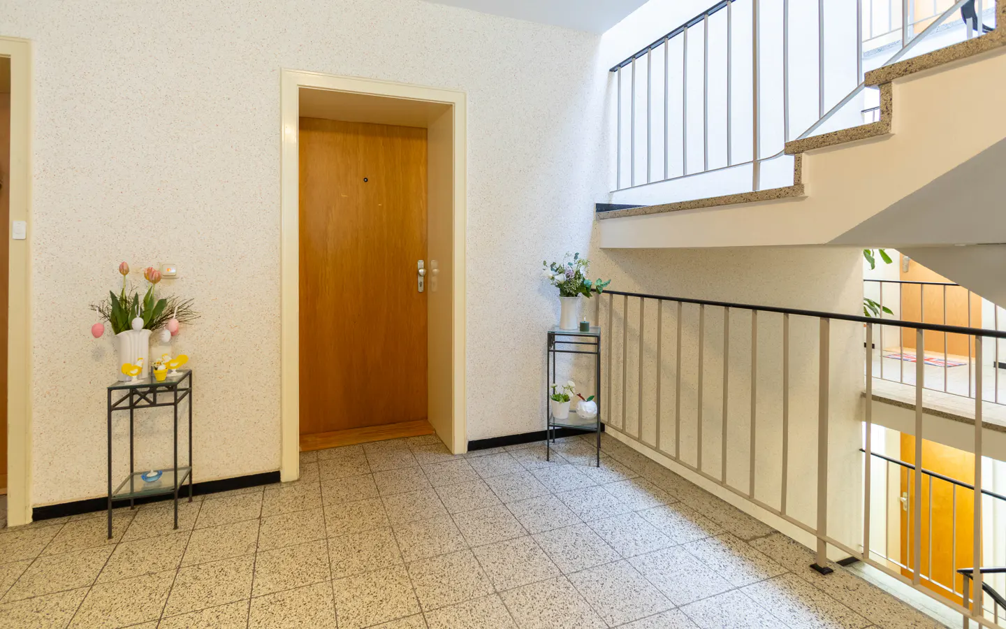 Hallway with beige tile floor, white walls, and a wooden door. A staircase with metal railings is on the right. Flowers decorate a small table.