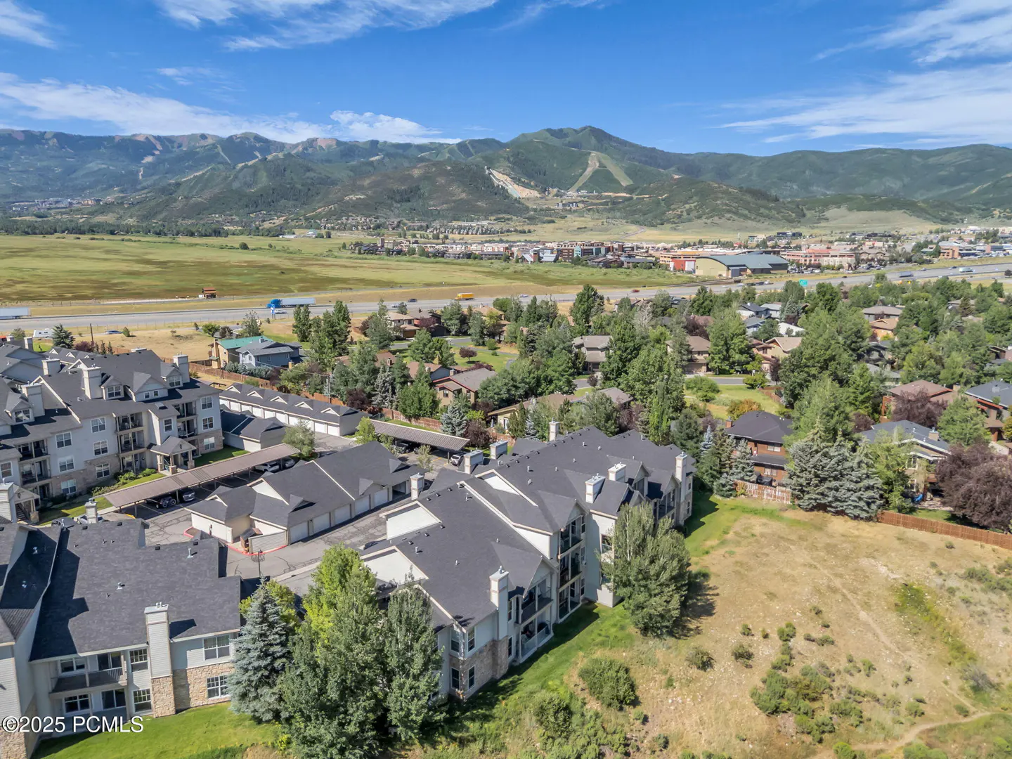 Aerial view of multi-story condos with dark roofs, green trees, and mountains in the background under a blue sky.