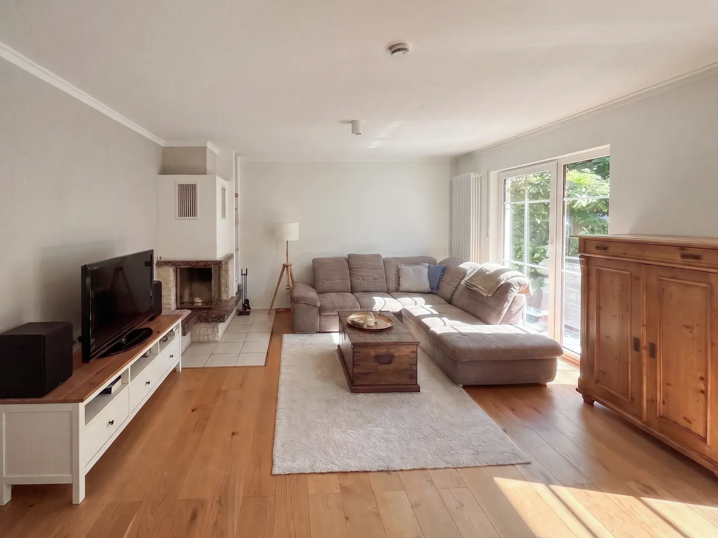 Bright living room with hardwood floors, a gray sectional sofa, a fireplace, and a TV on a white console. Sunlight streams in from a sliding glass door.