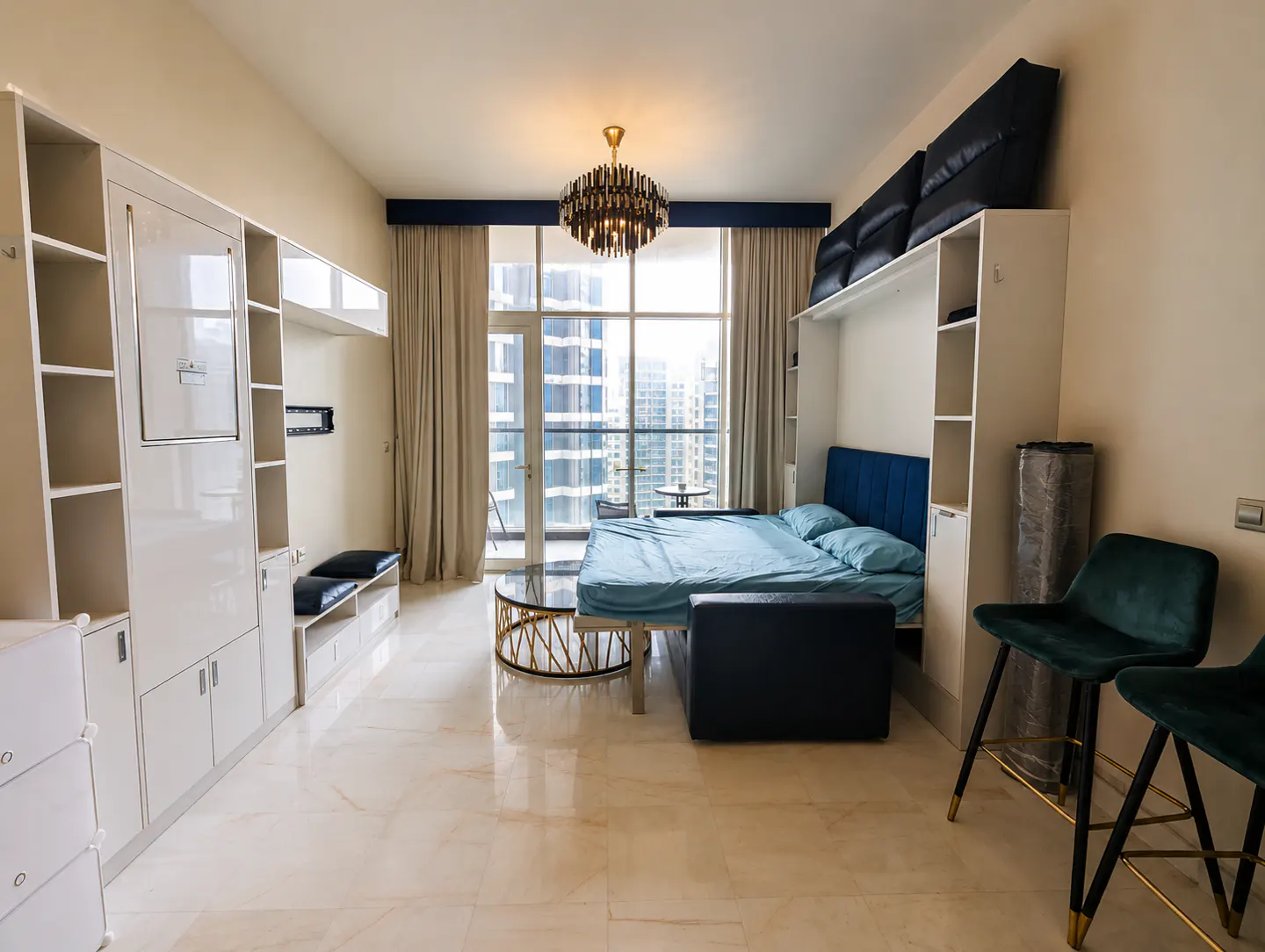 Apartment interior with a blue bed, white cabinets, and a city view. A modern chandelier hangs above the bed. Two green chairs are in the foreground.