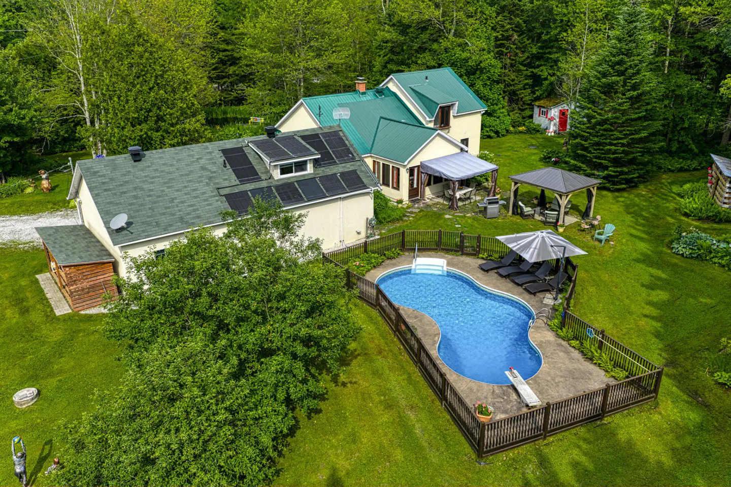 Aerial view of a yellow house with green roofs, solar panels, and a kidney-shaped pool surrounded by a wooden fence.